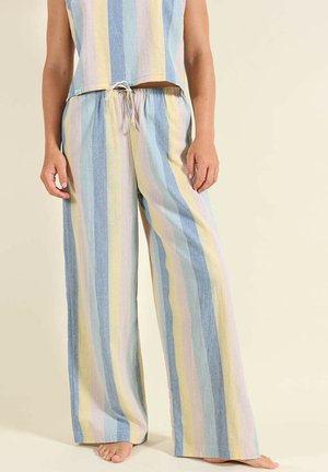 Femme portant un pantalon ample à rayures verticales pastel bleu, jaune et rose, debout pieds nus sur un fond uni.