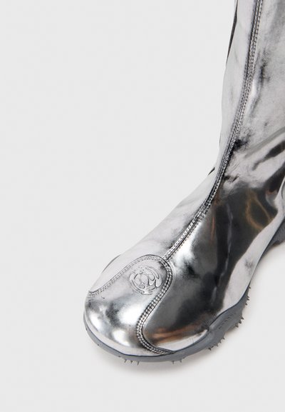 Stivale in metallo brillante argentato con suola in gomma nera, caratterizzato da piccoli spuntoni e un'emblema circolare cucito nella zona delle dita.