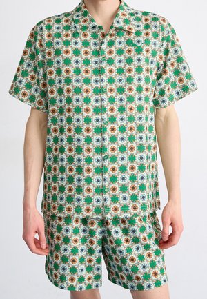 Homme portant une chemise à manches courtes avec boutons et un short assorti avec un motif géométrique vert, orange et bleu sur fond blanc.