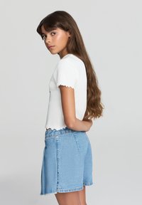 T-shirt bianco cropped con bordi a scallop, abbinato a pantaloni shorts in denim azzurro chiaro a vita alta, caratterizzati da spacchi laterali e orlo sfilacciato.