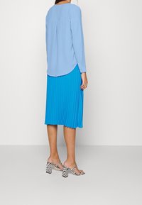 Haut bleu clair à manches longues avec un ourlet incurvé et des plis, associé à une jupe plissée bleue. Sandales à talons blocs à motifs noir et blanc.