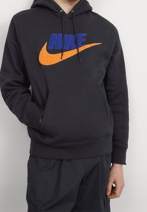Persoon draagt een zwarte hoodie met blauwe "NIKE" tekst en een oranje swoosh-logo, zwarte broek, staand met één hand in de zak.