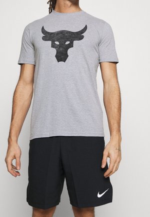 Mężczyzna ubrany w szary t-shirt z czarnym logo byka i czarne spodenki Nike z białym znakiem Swoosh na jednolitym tle.