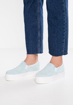 Zapatos slip-on de ante azul claro con una suela de plataforma blanca, que cuentan con paneles laterales elásticos y una textura suave, combinados con jeans azul oscuro.