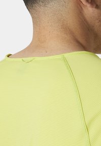 Detailní záběr na krátký rukáv, světle žlutou košili s ribbed texturou, s kulatým výstřihem a viditelnými švy.