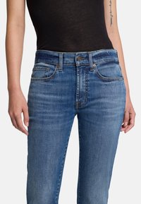 Niebieskie dżinsy z wysokim stanem, pięcioma kieszeniami i delikatnym efektem wyblaknięcia. Widoczne kontrastowe szwy oraz metalowe nity.