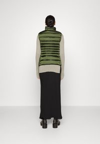 Grøn puffervest med vandret quiltning, høj krave, båret over en lys grå sweater med lange ærmer og en sort gulvlængde nederdel, iført sorte støvler.