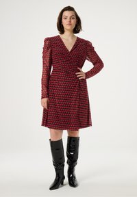 Rotes Kleid mit Herzmuster, langen transparenten Puffärmeln, Wickeldesign und einem ausgestellt Rock. Kombiniert mit schwarzen kniehohen Lackstiefeln.