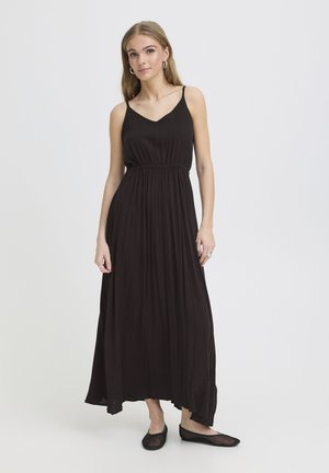 Joven con cabello largo, vestida con un vestido negro maxi sin mangas y zapatos planos negros, de pie contra un fondo claro y liso.