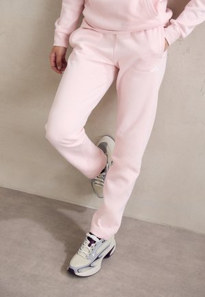 Pantalones de chándal rosa claro con una textura suave, con cintura ajustable y bolsillos laterales. Combinados con zapatillas deportivas grises y blancas.