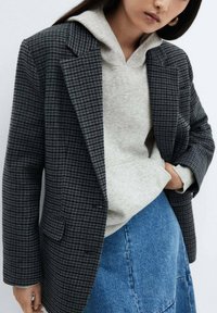 Grijze houndstooth blazer met notch-lapels, gedragen over een lichtgrijze hoodie, gecombineerd met een blauwe denim rok. Ontspannen pasvorm, gestructureerde stof.