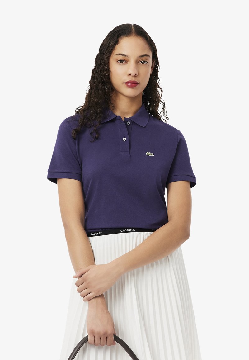 Camisa polo morada con un cuello acanalado y mangas cortas, que presenta un pequeño logotipo verde de Lacoste. Combinada con una falda blanca con pliegues.