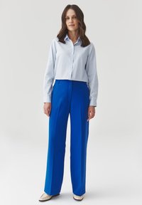 Camisa de botones azul claro combinada con pantalones de tiro alto y bien ajustados en azul brillante y zapatos color blanco roto, creando un look moderno y limpio.