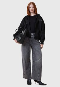 Sudadera negra cortada con logo, camiseta negra, vaqueros de pierna ancha grises con dobladillo deshilachado y botas de tacón negro de punta afilada. Bolsa tote negra en la mano.