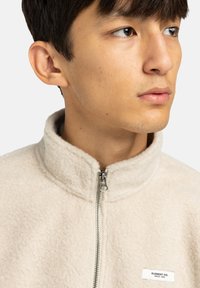 Beżowa kurtka polarowa z wysokim kołnierzem, zapinana na zamek z przodu, o teksturowanej powierzchni. Posiada małą metkę na piersi z informacjami o marce.