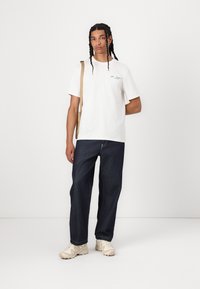 Hvid bomulds t-shirt med korte ærmer og blå grafisk detalje, parret med mørkeblå løstsiddende jeans og beige sneakers. Lysebrun crossbody taske.