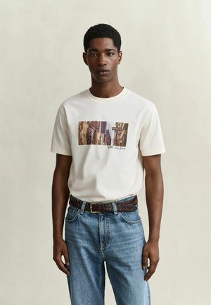 Jonge man die een wit T-shirt draagt met New York straatprint, blauwe spijkerbroek en gevlochten bruine riem, staand tegen een effen lichte achtergrond.