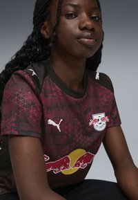 RB Leipzig trøje i mørkerød med geometriske mønstre. Har sorte accenter, hvid Puma-logo på skuldrene og Red Bull-emblemet på fronten.