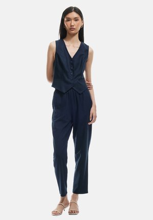 Navyblauwe op maat gemaakte gilet met een V-hals en zes knopen, gecombineerd met loszittende navy broek; gladde stof, minimalistisch ontwerp.