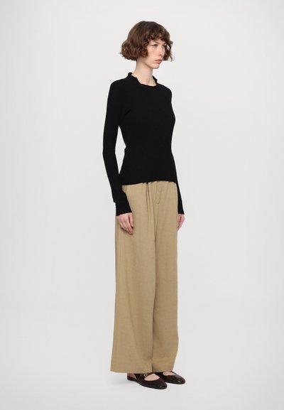 Black long-sleeve top with a scalloped neckline, paired with beige wide-leg pants. Model wearing dark brown flats. Minimalistic design.