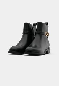 Botas de couro pretas até ao tornozelo, com bico arredondado, fecho lateral e uma fita de metal dourado. Superfície texturizada e salto baixo em bloco.