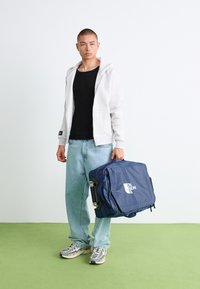 Sac de voyage bleu avec un logo, forme rectangulaire, poignées rembourrées et fermetures éclair. Porté avec un sweat à capuche gris clair et un jean en denim sur un sol vert.