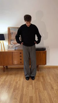 Νεαρός άνδρας με μαύρο πουλόβερ και γκρι παντελόνι στέκεται με τα χέρια στις τσέπες σε δωμάτιο με ξύλινο πάτωμα και έπιπλο μέσης εποχής.