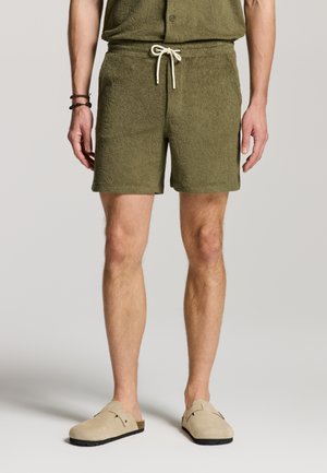 Mand iført teksturerede olivengrønne shorts med snøre, matchende skjorte, beige slip-on sko og sorte perlearmbånd på venstre håndled.