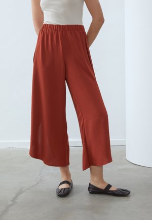 Woman wearing loose, rust-colored wide-leg pants with an elastic waistband, white tank top, and black ballet flats standing on a concrete floor.