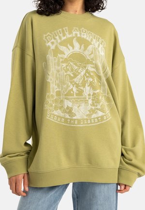 Persoon draagt een oversized olijfgroene trui met woestijnthema-afbeelding met cactussen, bergen, zon en tekst "Billabong 1973."