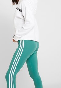 Kvinna i turkosa leggings med vita ränder och en vit Adidas-hoodie mot en enfärgad bakgrund, sedd från sidan midjan ner.