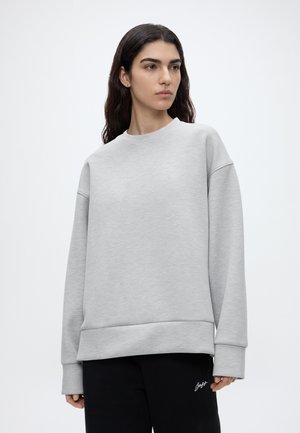 Jeune femme aux longs cheveux foncés porte un sweat-shirt oversize gris clair uni et un pantalon noir avec un logo en écriture blanche.
