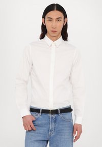 White long-sleeve button-up shirt with a pointed collar, paired with light blue denim jeans and a black belt. Smooth fabric, minimalistic design.