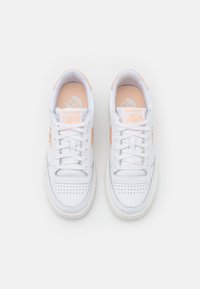 Sneaker in pelle bianca con accenti in arancione pastello. Caratteristiche includono una punta perforata, lacci piatti e una soletta imbottita. Loghi sulla linguetta e sul lato.