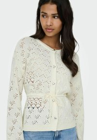 Cardigan en tricot crème avec motif ajouré, manches longues, boutons argentés en forme de cœur et taille à cordon. Porté sur un jean.