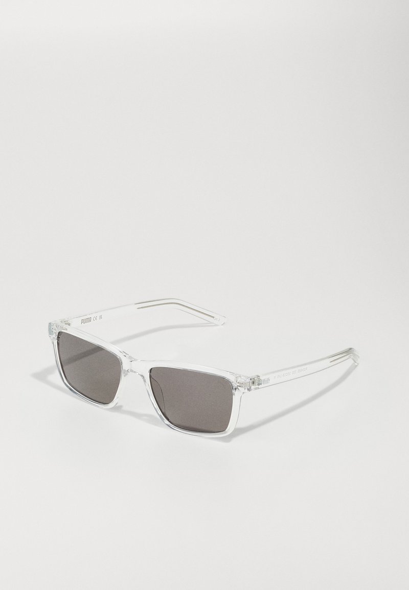 Lunettes de soleil en plastique transparent avec des verres gris foncé, de forme rectangulaire et avec des branches fines. La branche gauche présente le logo de la marque imprimé en blanc.