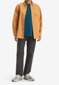 Camicia button-up arancione chiaro sopra una maglietta teal, abbinata a jeans scuri e sneakers grigie da ginnastica. Due tasche visibili sulla camicia.