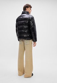 Μαύρο μπουφάν με γυαλιστερή επιφάνεια, διαθέτει οριζόντια καπιτονέ τμήματα και ψηλό γιακά. Συνδυάζεται με μπεζ παντελόνι σε φαρδιά γραμμή.