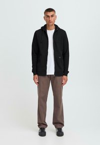 Haut à capuche zippé noir avec cordons de serrage, porté sur une chemise blanche, associé à un pantalon marron et des baskets noires. Design simple et fonctionnel.