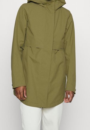 Persoon draagt een olijfgroene jas met capuchon, lange mouwen en een verborgen sluiting aan de voorkant, gecombineerd met een witte broek.