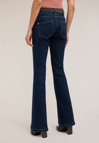 Tummansiniset leveälahkeiset farkut, joissa on keskikorkea vyötärö, sileä pinta, kaksi takataskua ja ruskea vyö. Neliönmuotoiset korokepohjaiset kengät.