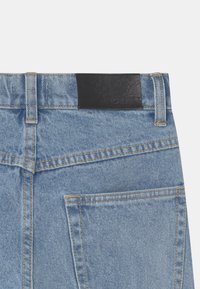 Lysblå denimbukser med en sort læderbelægning på taljen. Har synlige sømme og en baglomme på højre side.