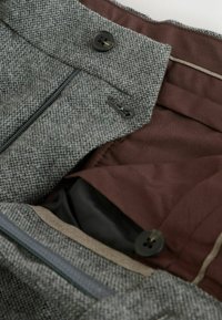 Gray tweed fabric with a herringbone pattern, featuring a dark button, zipper, and brown lining. Close-up of pockets and stitching details.