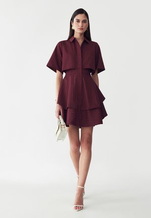 Donna con lunghi capelli scuri indossa un vestito corto bordeaux a righe sottili e tacchi bianchi, porta una piccola borsa bianca, in piedi su uno sfondo semplice.