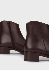 Botas de tobillo de cuero marrón con un acabado suave, con cremallera lateral y un tacón bajo en bloque. El nombre de la marca está grabado en el tacón.