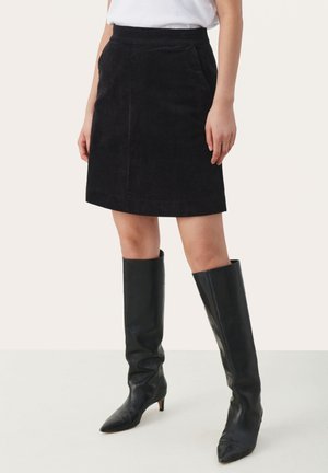 Jupe en velours côtelé noire avec une silhouette droite, dotée de poches sur les côtés et d'une ceinture, associée à des bottes en cuir noir jusqu'aux genoux.