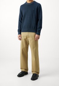 Dunkelblauer Strickpullover mit geripptem Halsausschnitt und Bündchen, kombiniert mit beigefarbenen Chinos und schwarzen Schuhen, vor einem weißen Hintergrund stehend.