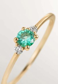 Anillo de oro con una piedra redonda central de color teal, flanqueada por dos piedras claras más pequeñas, con engastes de garras redondeadas y una banda lisa.