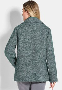 Femme aux longs cheveux blonds portant un manteau en laine à motif chevron vert et blanc, se tenant de dos avec une main sur la hanche.
