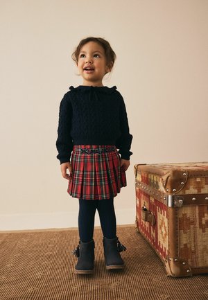 Pull en maille bleu marine avec col, jupe plissée écossaise rouge à carreaux, collants côtelés bleu marine, et bottines noires avec pompons, debout sur un tapis texturé.